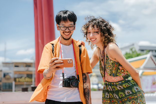 Casal sorridente olhando para um smartphone ao ar livre, com um fundo colorido e vibrante

