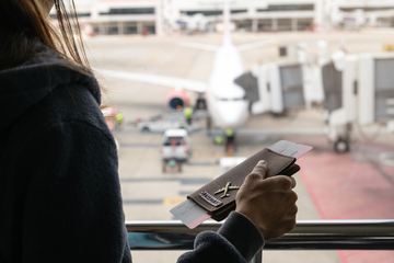 Pessoa segurando bilhetes de embarque em um aeroporto, com vista para um avião e equipe de solo ao fundo.
