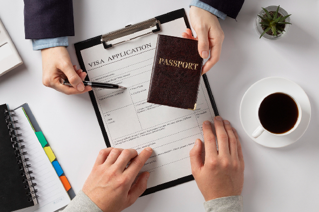 Mãos segurando um passaporte e apontando para um formulário de solicitação de visto em uma prancheta, com uma xícara de café e uma planta ao fundo.
 Mãos segurando um passaporte e apontando para um formulário de solicitação de visto em uma prancheta, com uma xícara de café e uma planta ao fundo.
